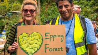 Two people holding a sign with a leafy heart and 'Epping Forest' in a wooded area.