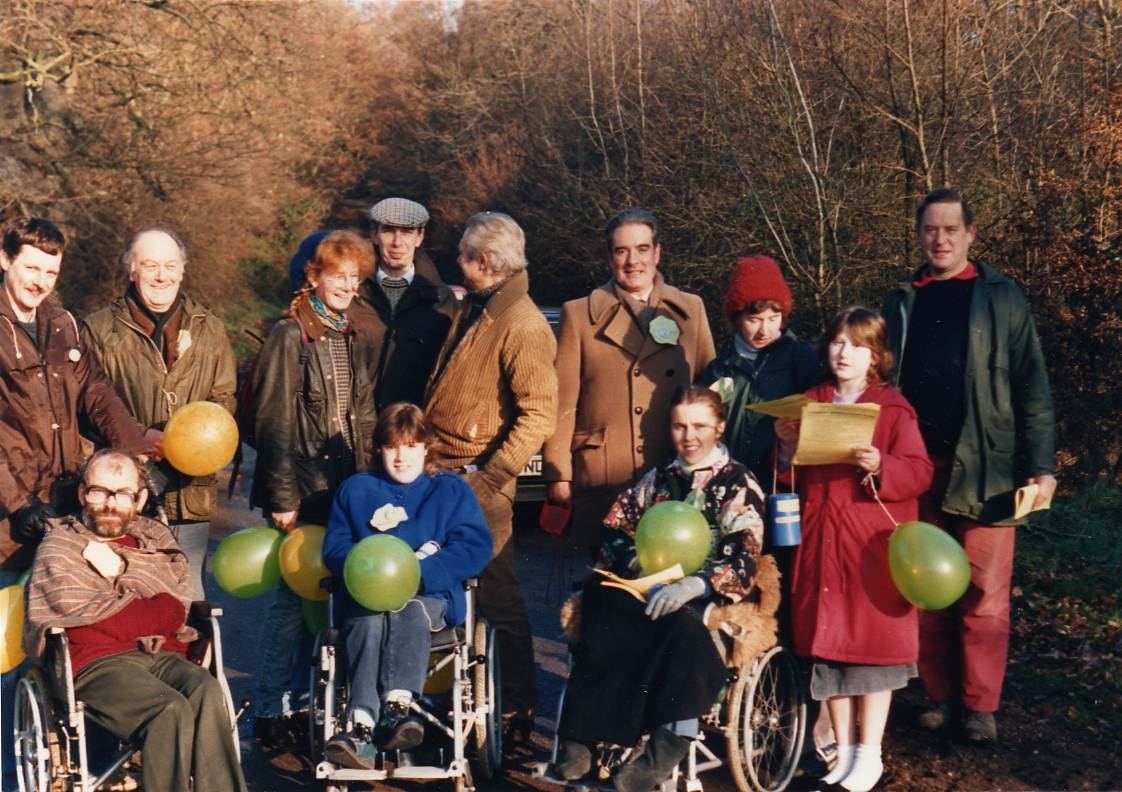 Community gathering of about a dozen people gathered outdoors on a sunlit pathway bordered by leafless trees. They wore warm coats, hats, suggesting it was a cold day. The group is positioned across the width of the path, facing toward the camera. Several individuals stand while three people sit in wheelchairs at the front, some holding balloons. 