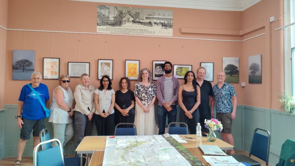 Group photo of ten participants of EFHT's first People's Forest Panel standing in Chingford community hall behind tables displaying a large map of Epping Forest, papers and a vase of flowers. Framed artwork about the Forest hangs on the walls, and large windows bring natural light into the room. 