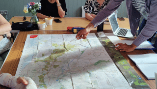 People gathered around a large map of Epping Forest spread across a table. One person points to a location on the map while others sit nearby. Cups, a vase of flowers, a laptop and papers are visible on the table, suggesting a collaborative discussions session.